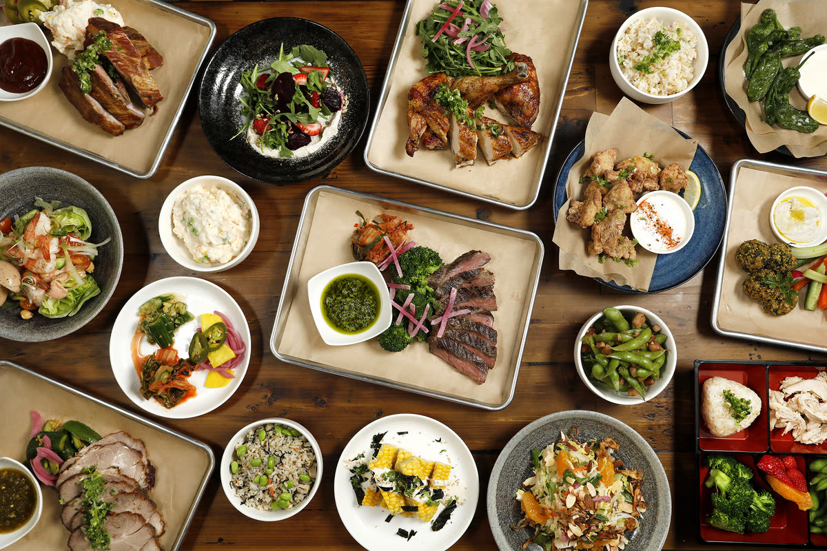 A wooden table covered with sharing trays — rotisserie chicken, sliced steak with chimichurri, ribs, salads, rice bowls and pickled vegetables.